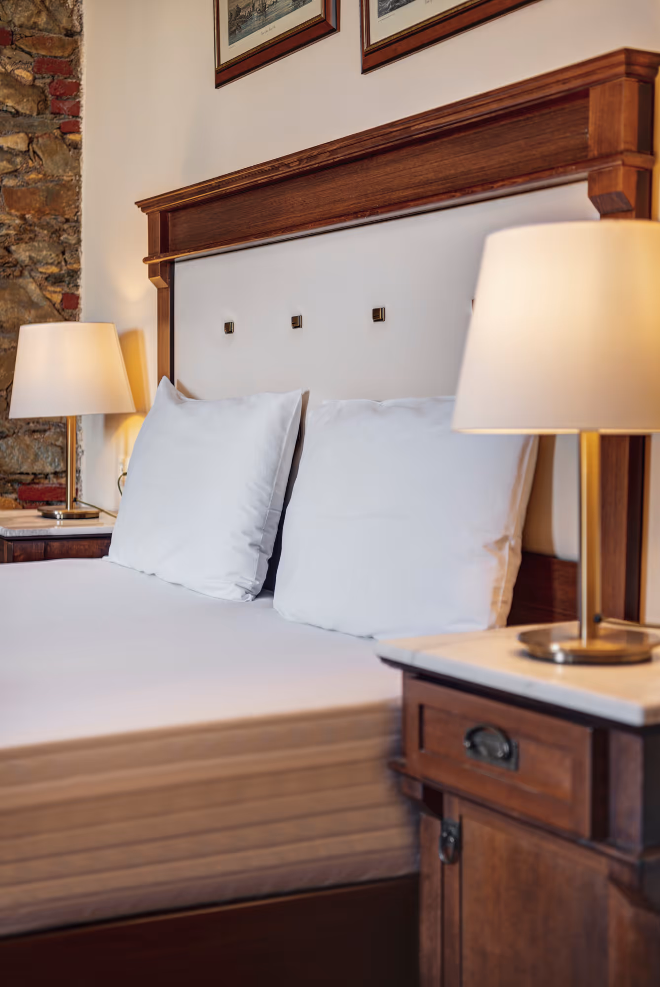 Close-up of a bed with white pillows and sheets, wooden headboard, bedside tables with lamps, and exposed stone wall in the background.