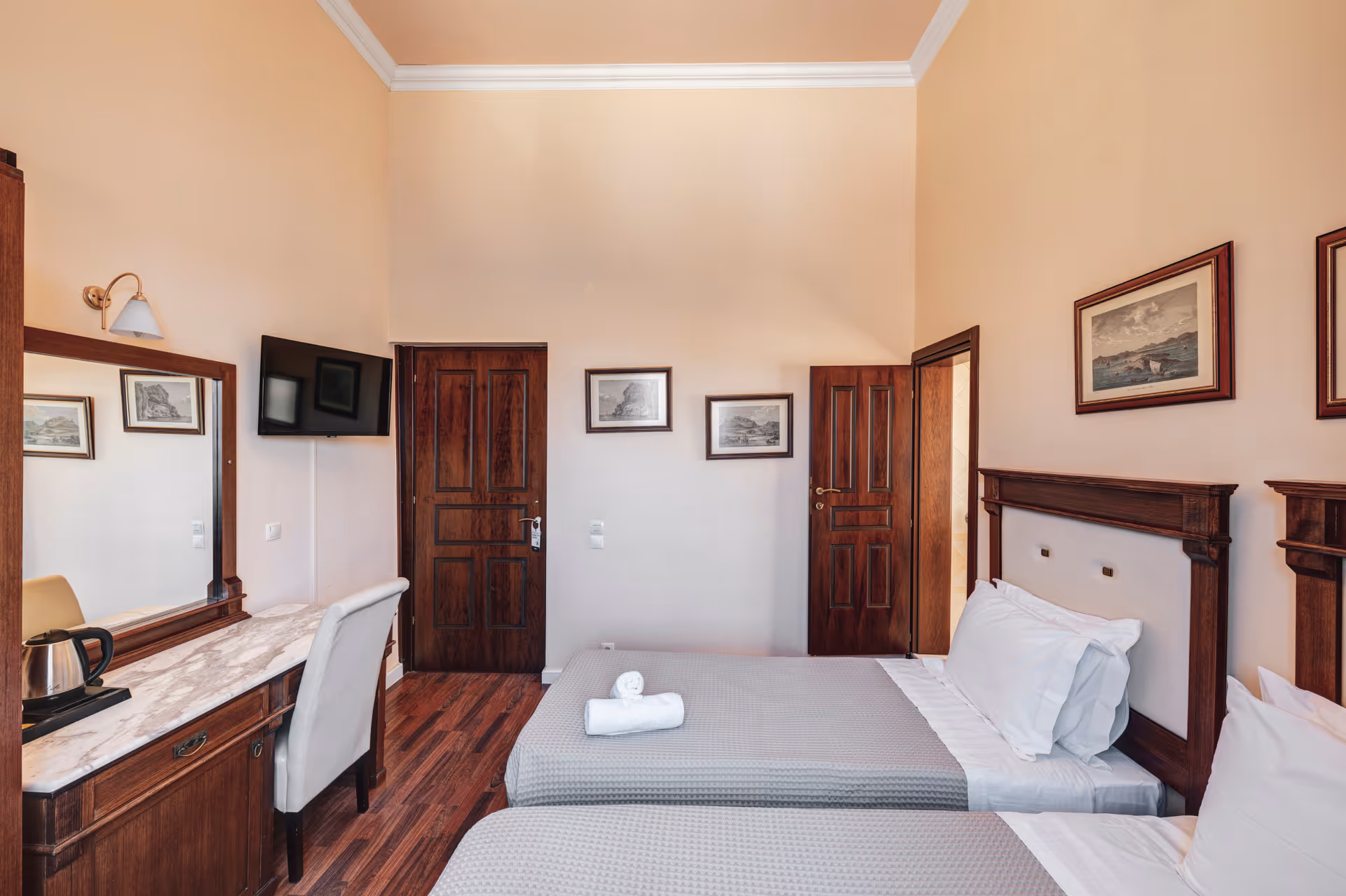 Hotel room with two single beds with gray bedspreads, wooden headboards, framed artwork on beige walls, a wooden desk with marble top, a white chair, and a wall-mounted TV.