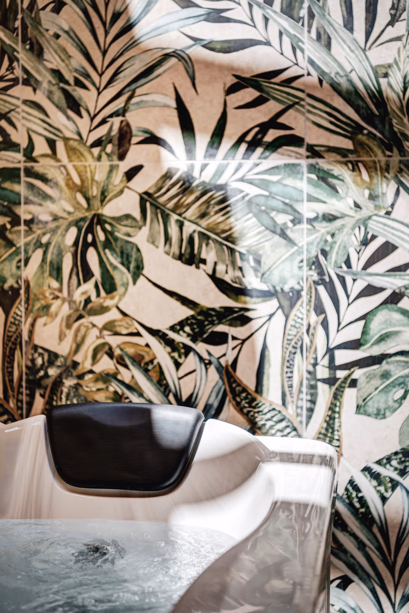 Whirlpool tub with black headrest in front of a wall covered with large green tropical leaf wallpaper.