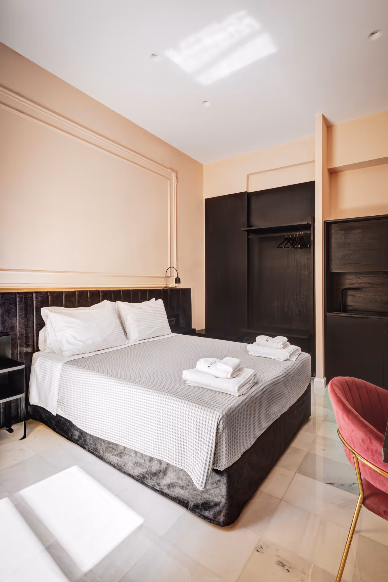 Modern hotel room with a neatly made double bed, white linens, two sets of folded towels, black open wardrobe, and a red velvet chair.