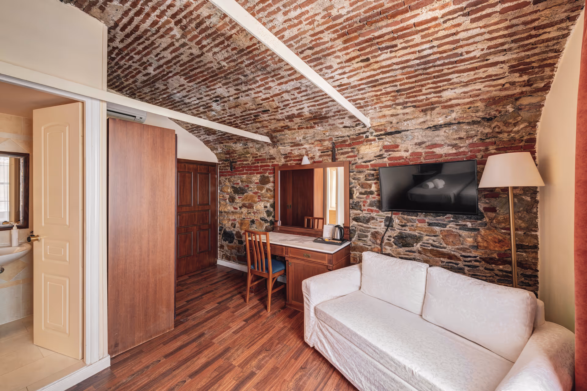 Cozy room with a curved exposed brick ceiling, wooden floor, white sofa, wall-mounted TV, wooden desk with mirror, and a door leading to a bathroom.
