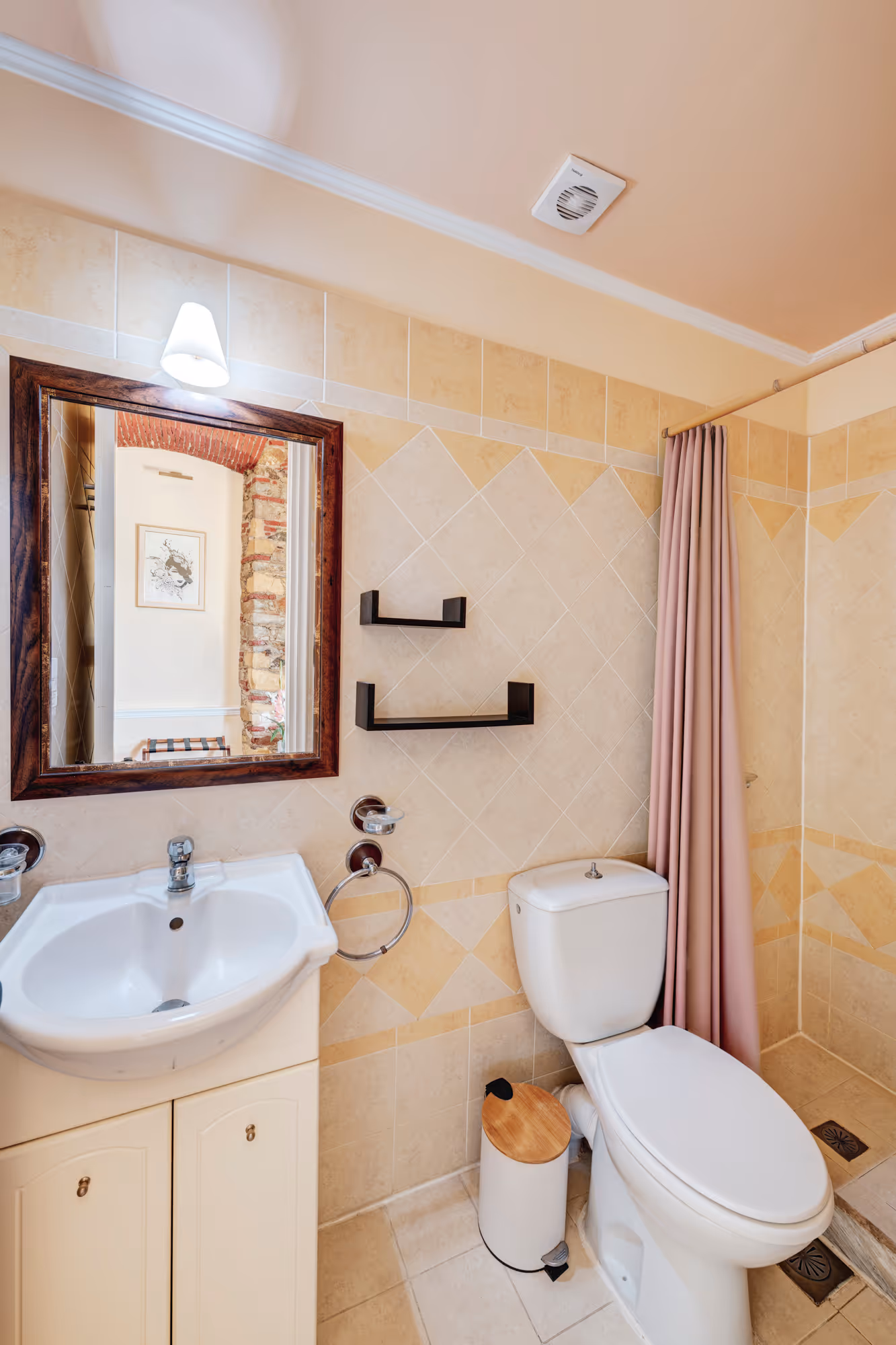 Small bathroom with white sink, wooden-framed mirror, white toilet, pink shower curtain, and beige tiled walls.