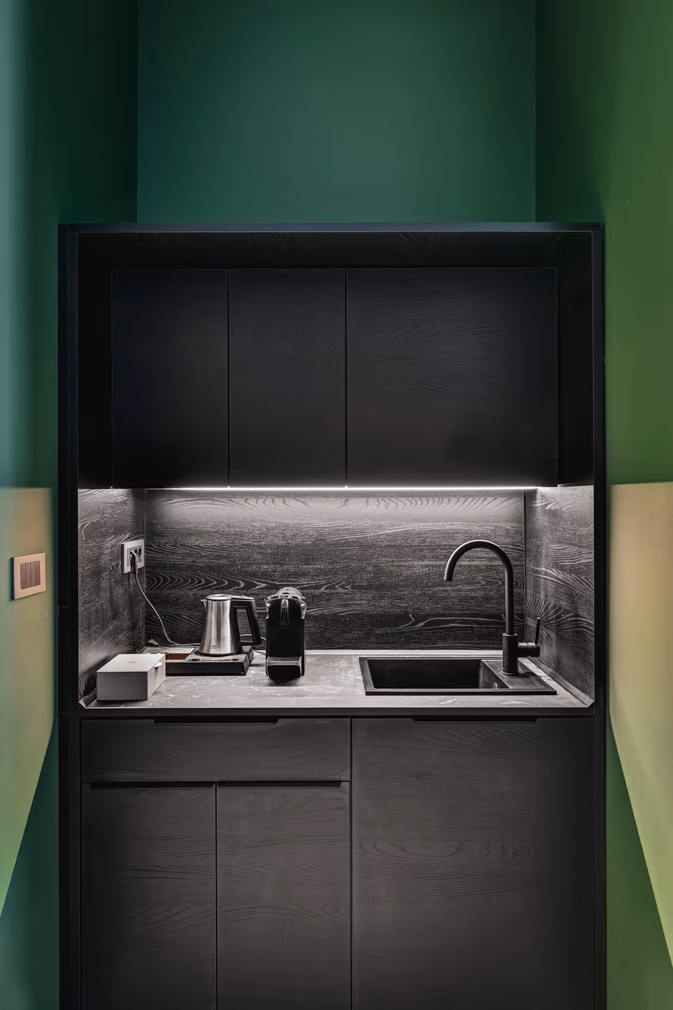 Compact modern kitchenette with black cabinets, black sink and faucet, electric kettle, and coffee machine, against green walls.