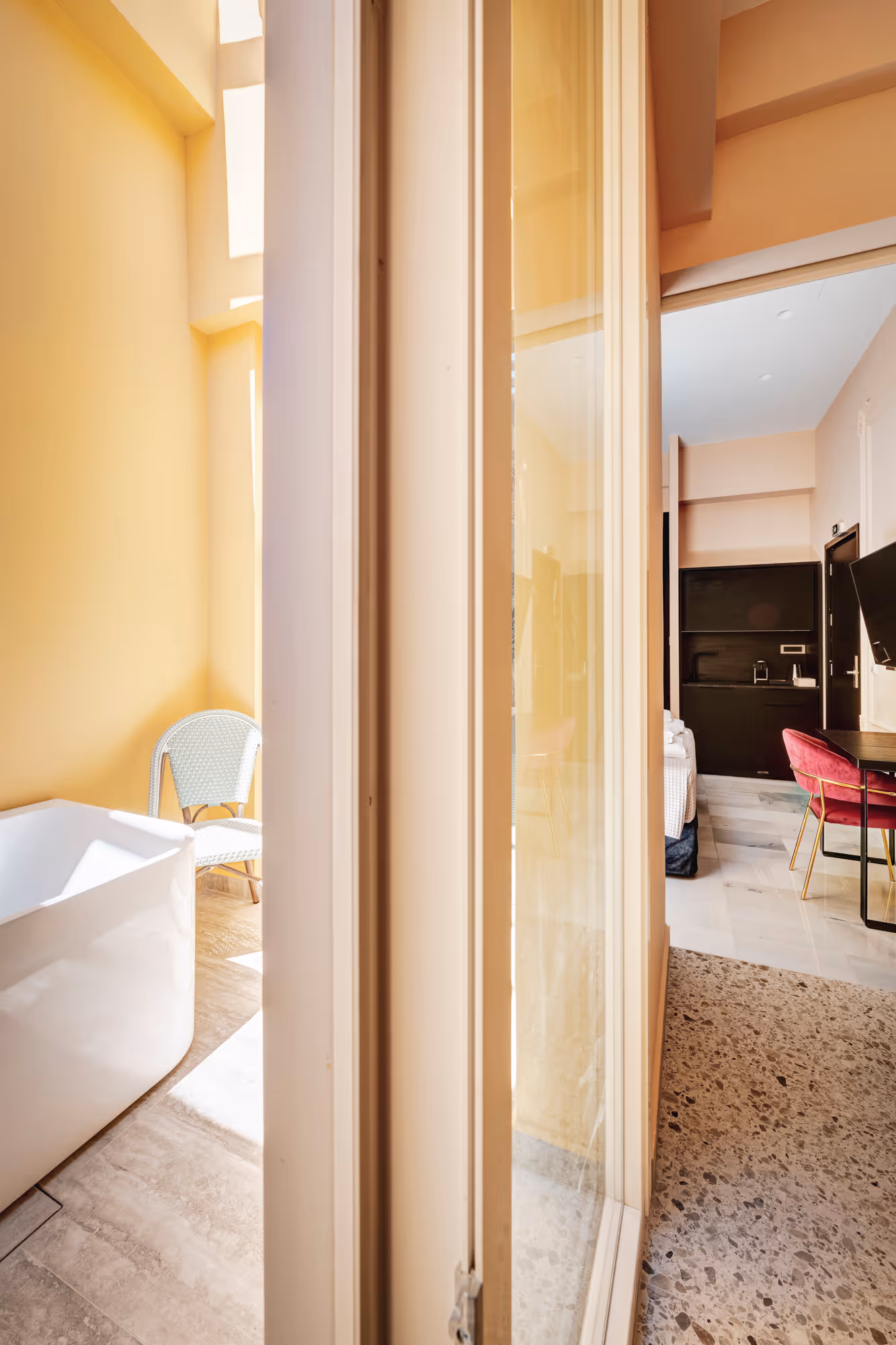 View through glass door of a room with terrazzo floor, pink chair, wall-mounted TV, kitchenette, and a yellow-painted balcony with white bathtub and chair.