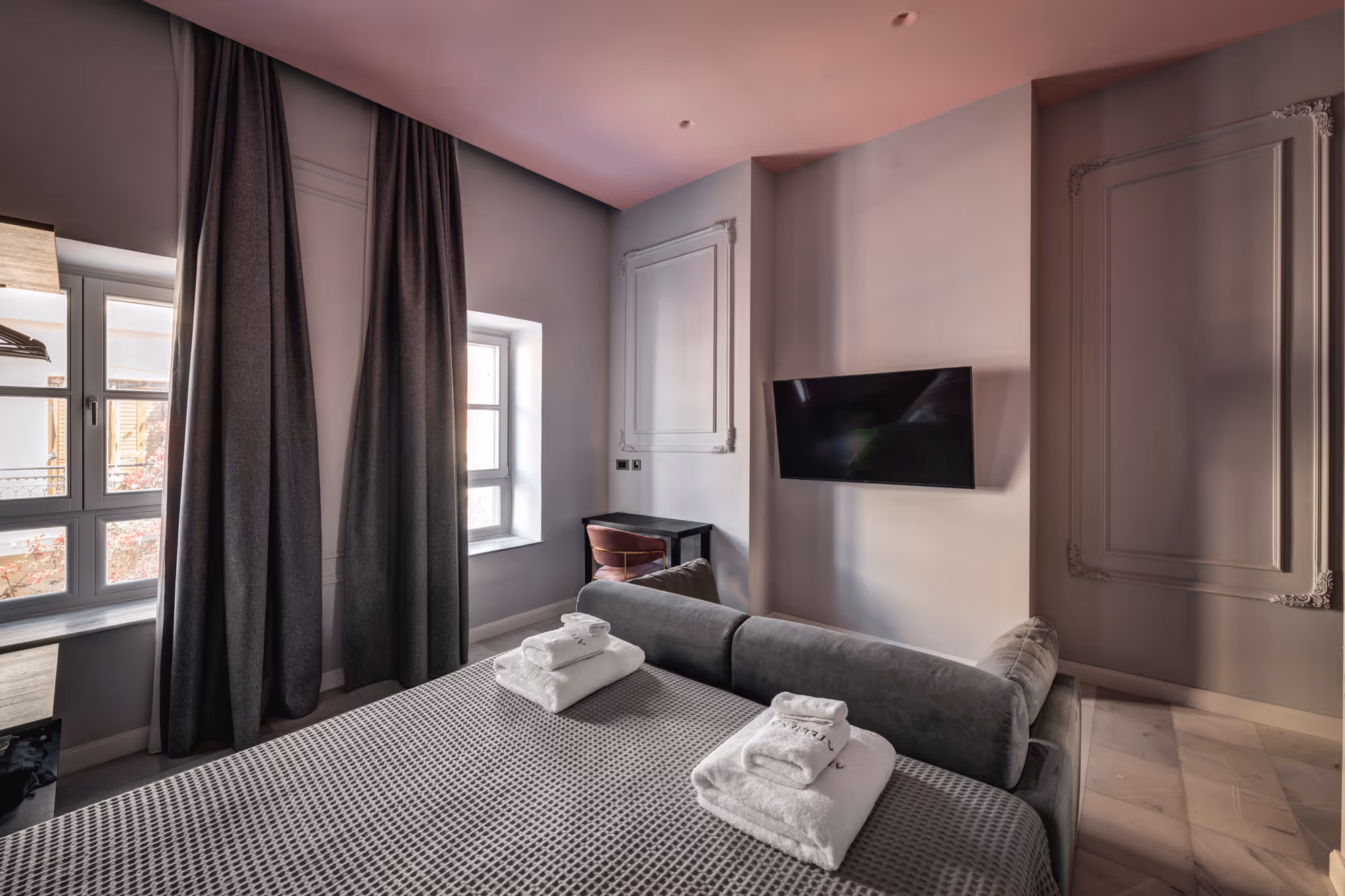 Modern hotel room with a bed covered in gray waffle-texture bedding, two sets of white folded towels, a gray couch at the foot of the bed, a wall-mounted flat-screen TV, and a small black desk with a chair near two windows with dark curtains.