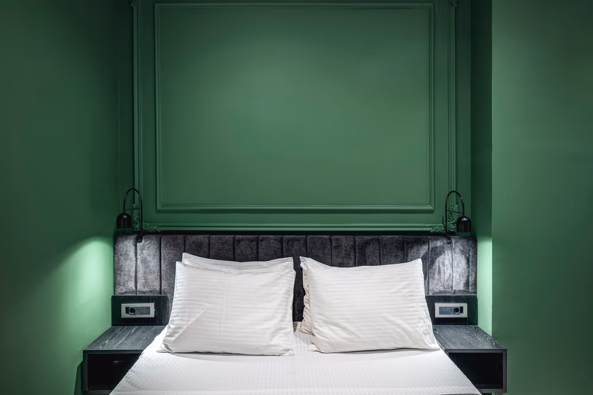 Modern bed with white pillows and sheets, dark upholstered headboard, green walls, and black bedside tables with small lamps.