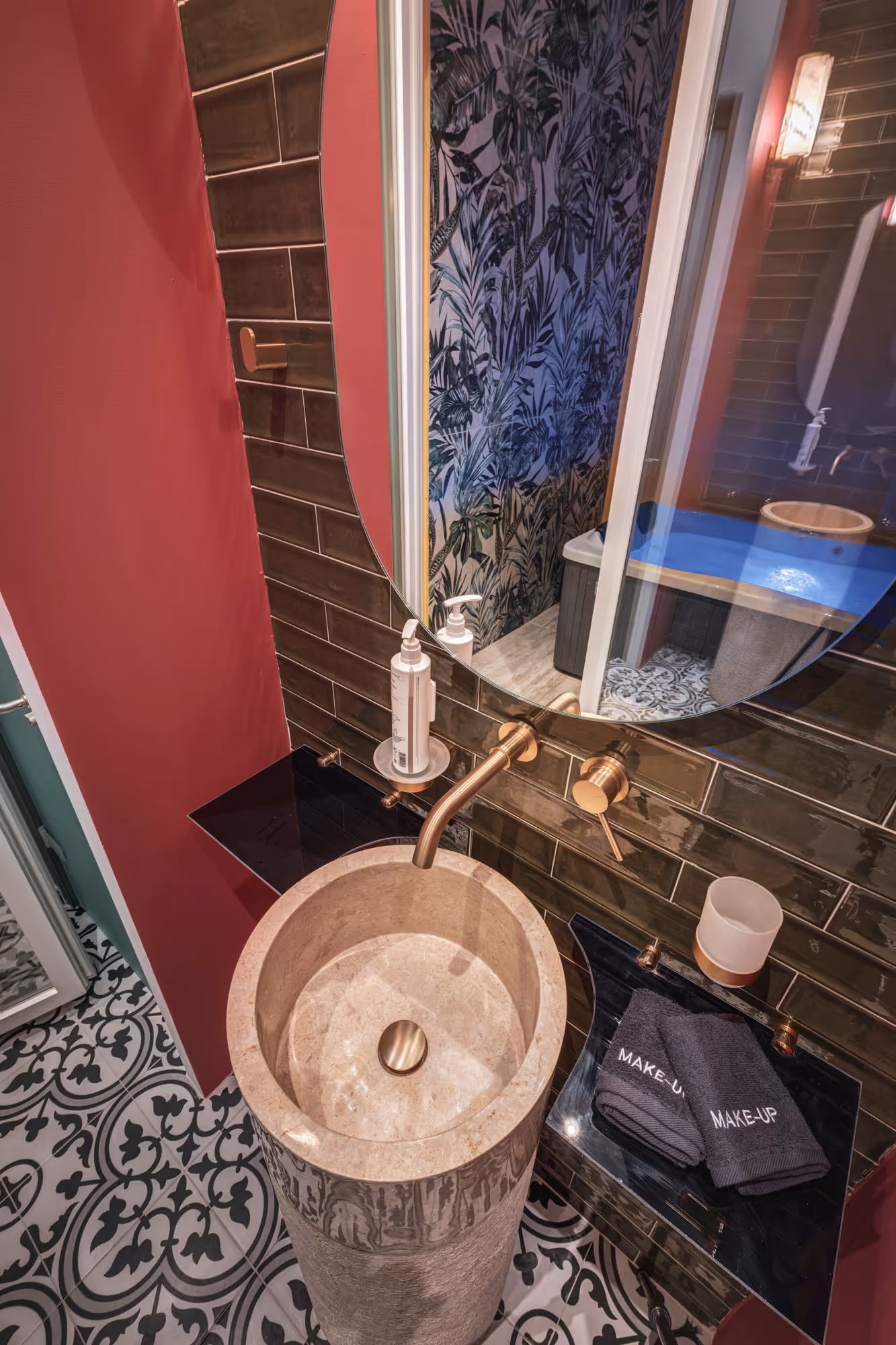 Stylish bathroom corner with a round stone pedestal sink, gold faucet, round mirror, black makeup towels, and patterned floor tiles.