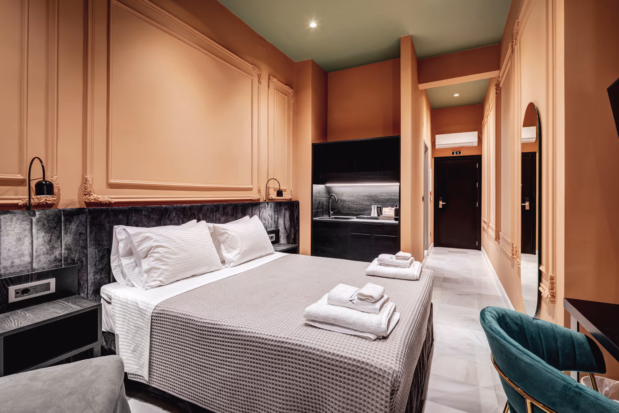 Modern hotel room with a large bed, gray bedspread, white pillows, folded towels, a kitchenette with black cabinets, a green velvet chair, and peach-colored walls.