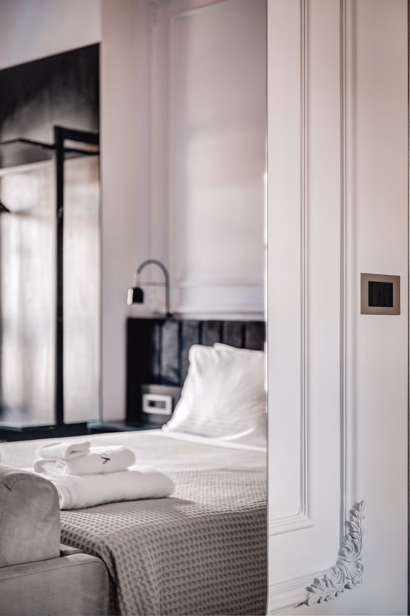 Modern bedroom with a neatly made bed covered with a gray textured blanket, white pillows, folded white towels on top, a black headboard, and white paneled wall with decorative molding.