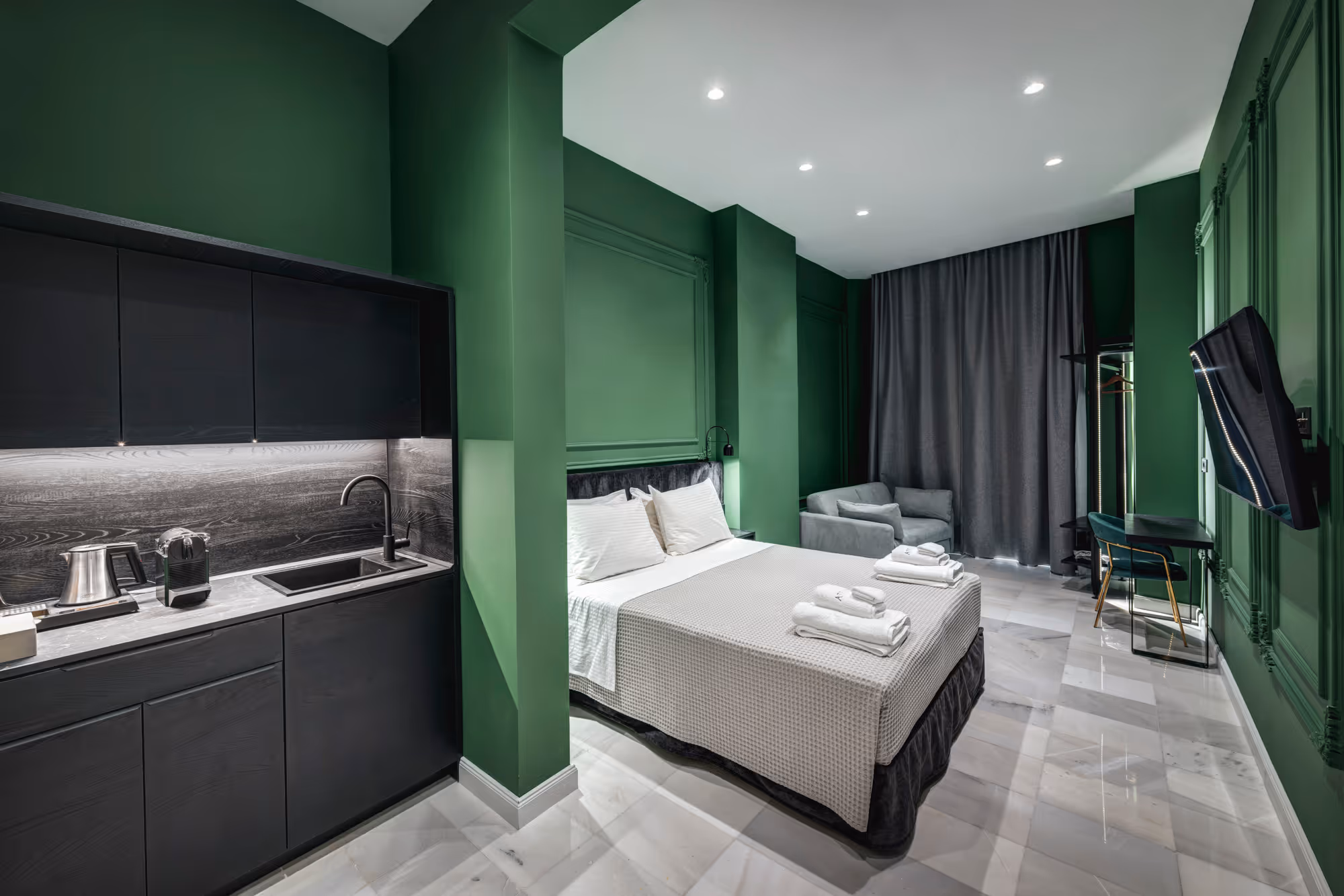 Modern studio apartment with green walls, kitchenette, double bed with folded towels, gray armchair, and wall-mounted TV.