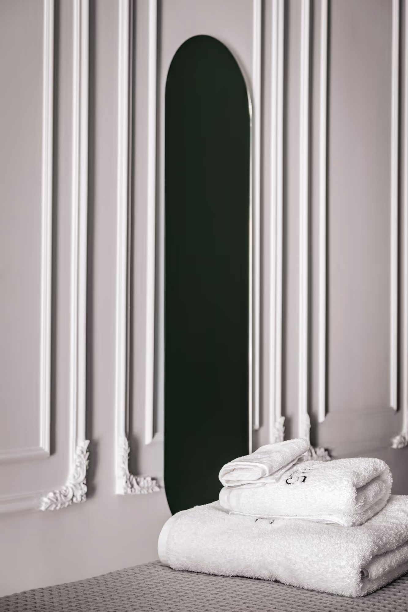 Stack of three white folded towels on a textured gray surface with a tall, narrow, dark green mirror against an ornate white paneled wall.