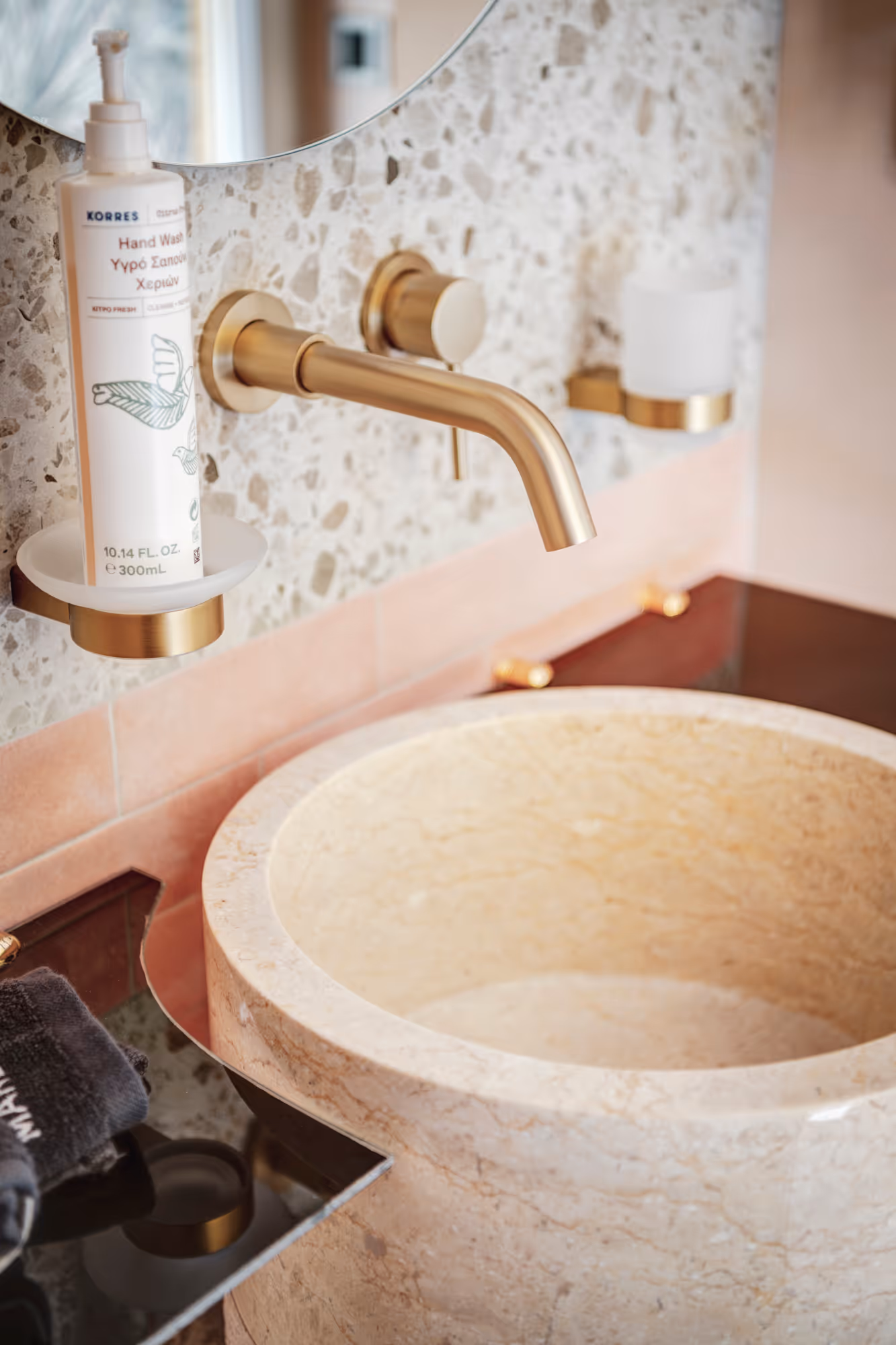 Close-up of a beige marble sink with a gold wall-mounted faucet and a bottle of Korres hand wash on a gold holder against a speckled wall.