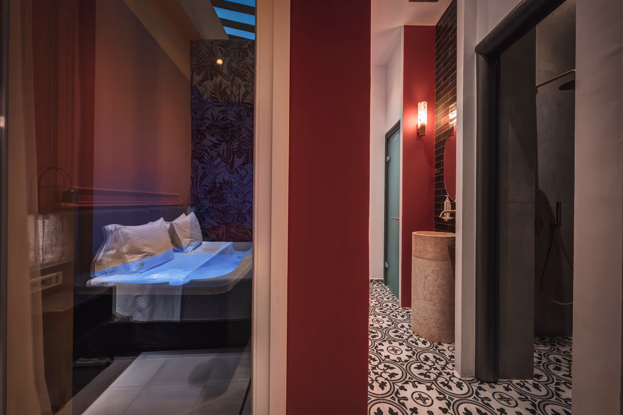 View of a modern hotel room with a blue-lit bed behind glass and a bathroom hallway with patterned floor tiles and red walls.