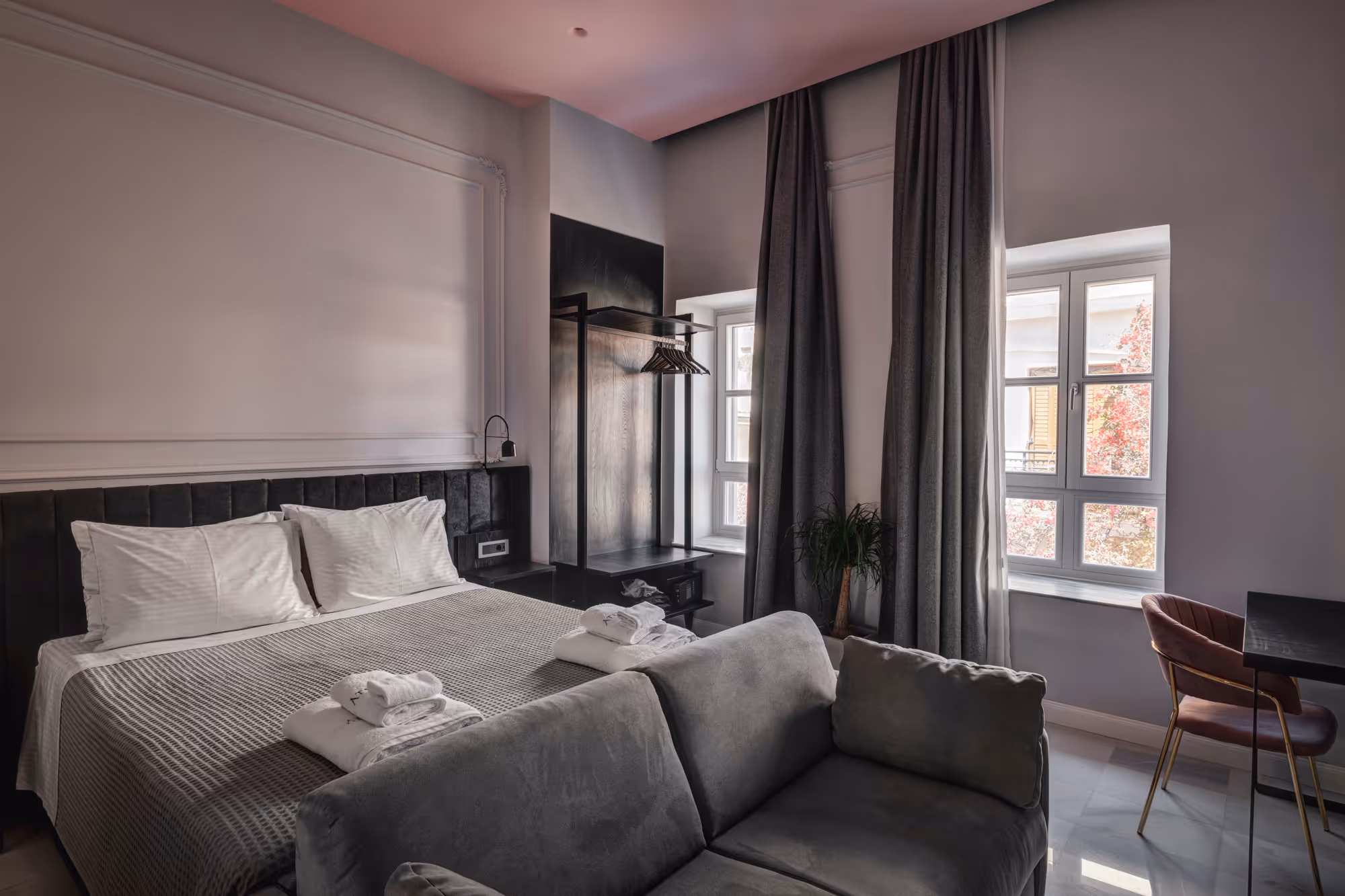 Modern hotel room with a double bed, gray sofa, window with curtains, and a small desk with a chair.