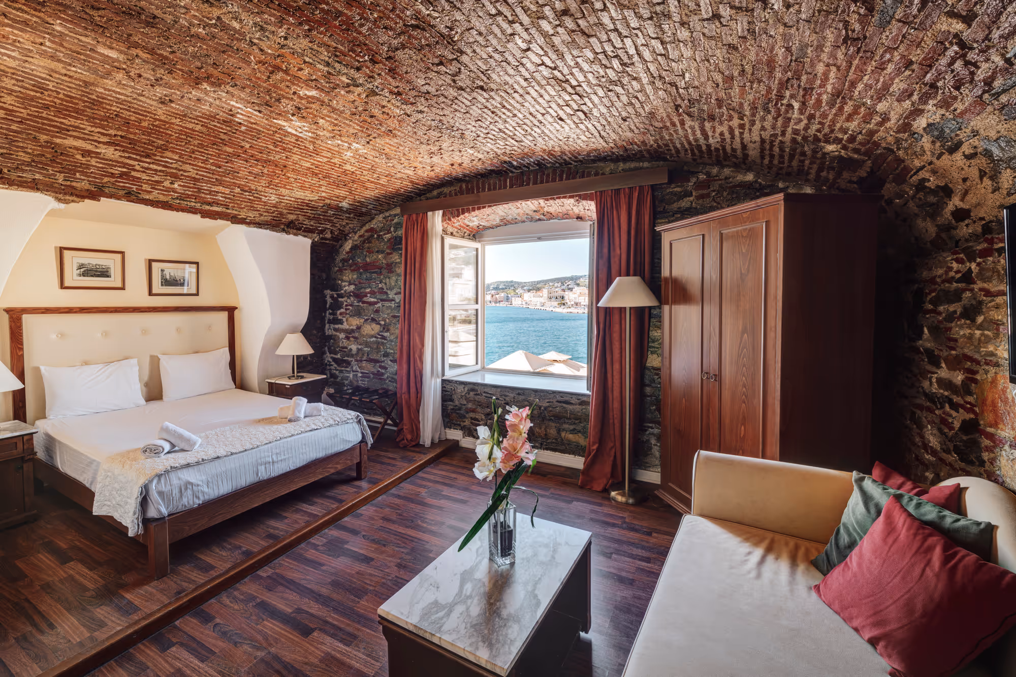 Cozy bedroom with arched brick ceiling, bed with white linens, wood wardrobe, sofa with colorful pillows, and window showing sea view.
