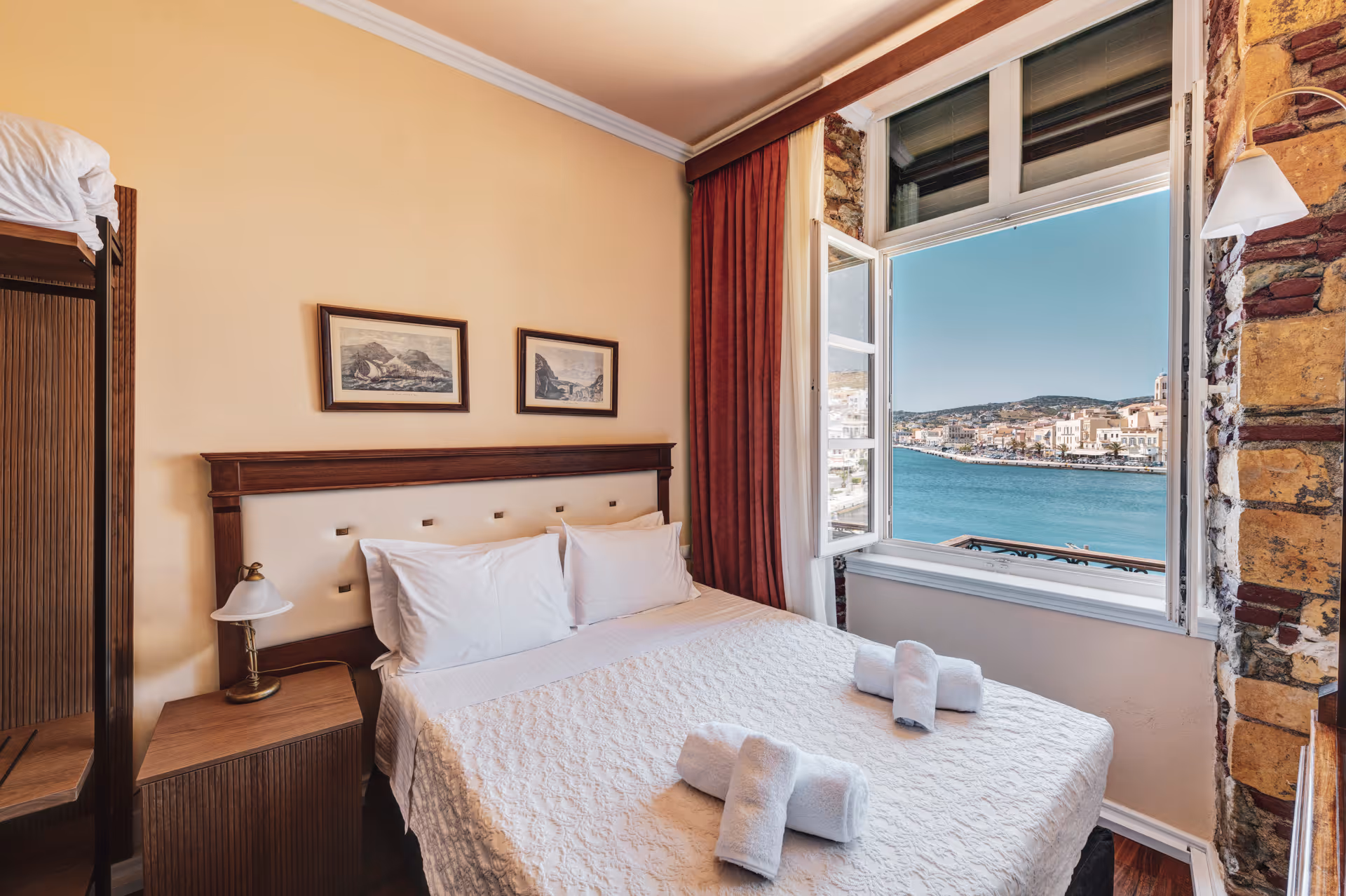 Cozy bedroom with a double bed, white linens, rolled towels, wooden furniture, and an open window overlooking a waterfront town.