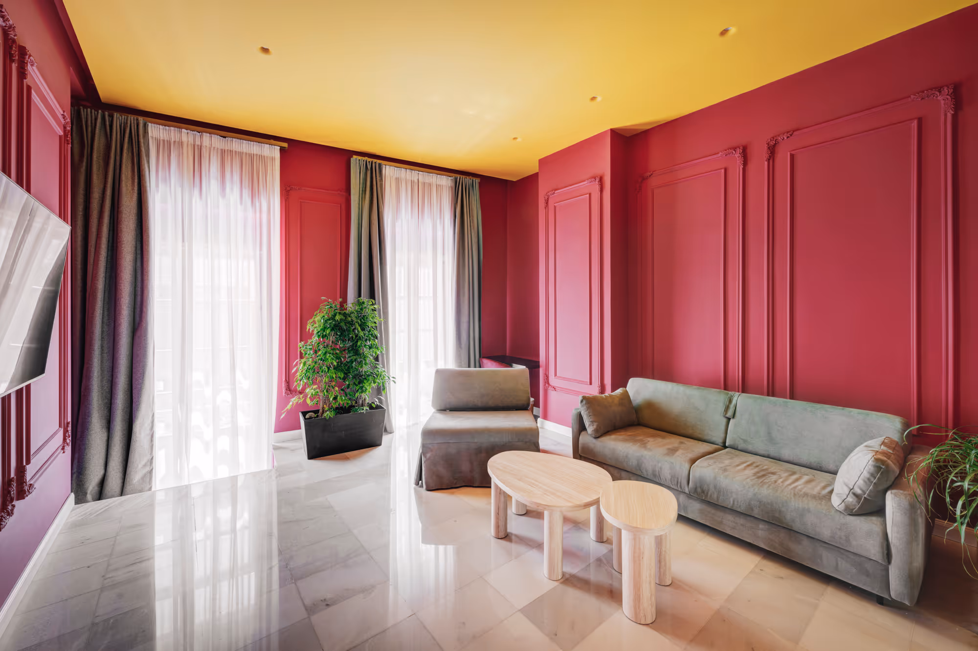 Modern living room with a gray sofa, matching chair, two wooden coffee tables, green plants, and magenta walls with decorative molding under a yellow ceiling.