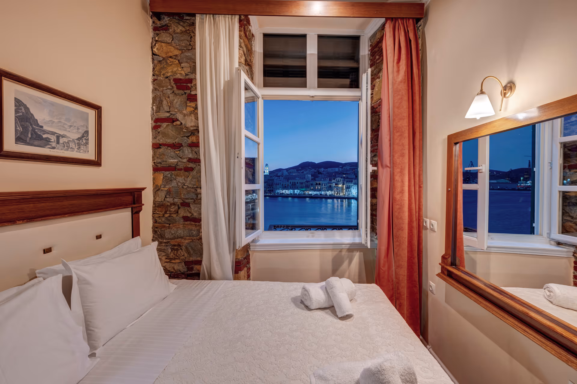 Cozy bedroom with white bedding, open window with a view of waterfront buildings at dusk, stone wall, and soft lighting.