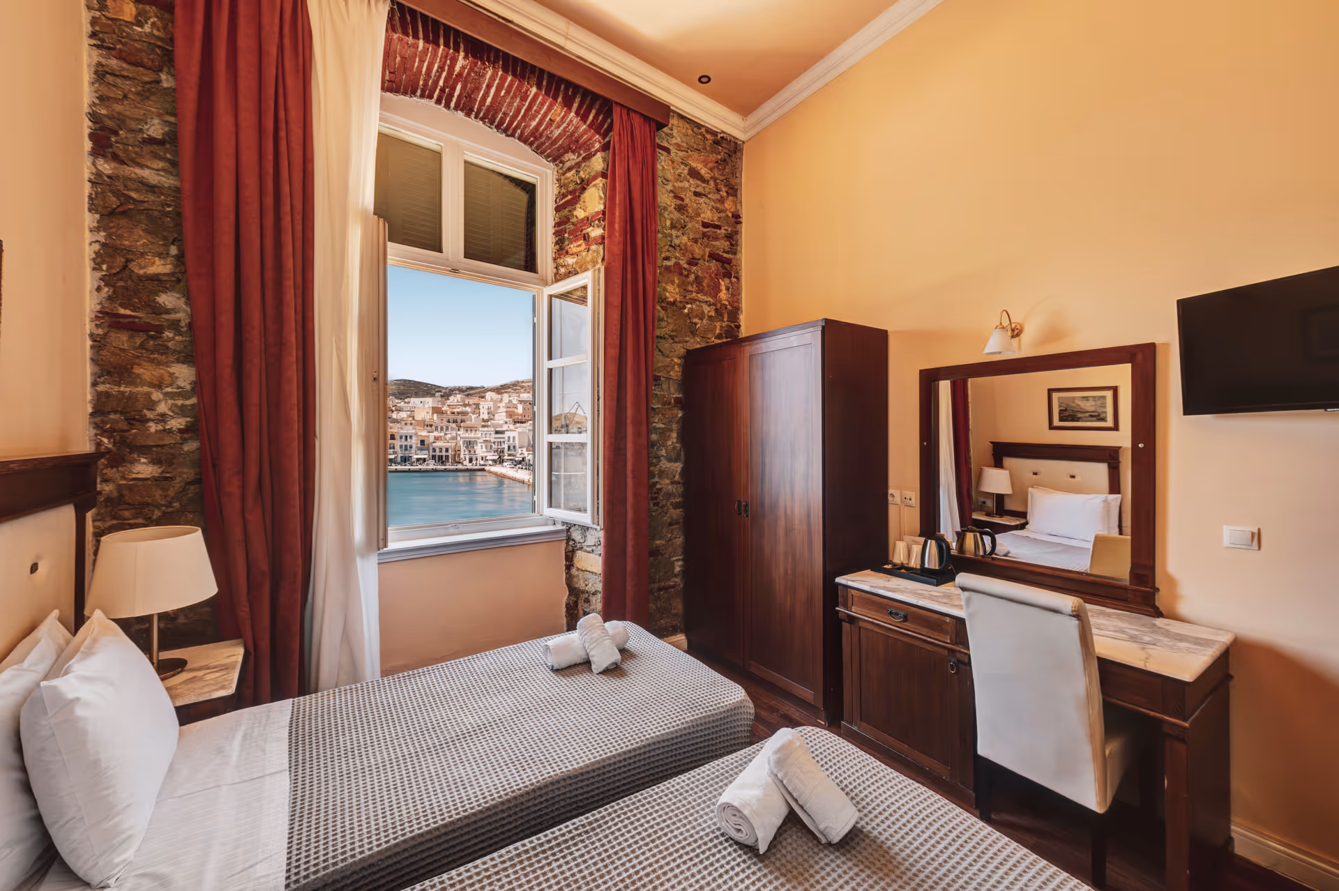 Hotel room with two single beds, each with white pillows and towels, a wooden dresser with a mirror and chair, a window showing a seaside town view.