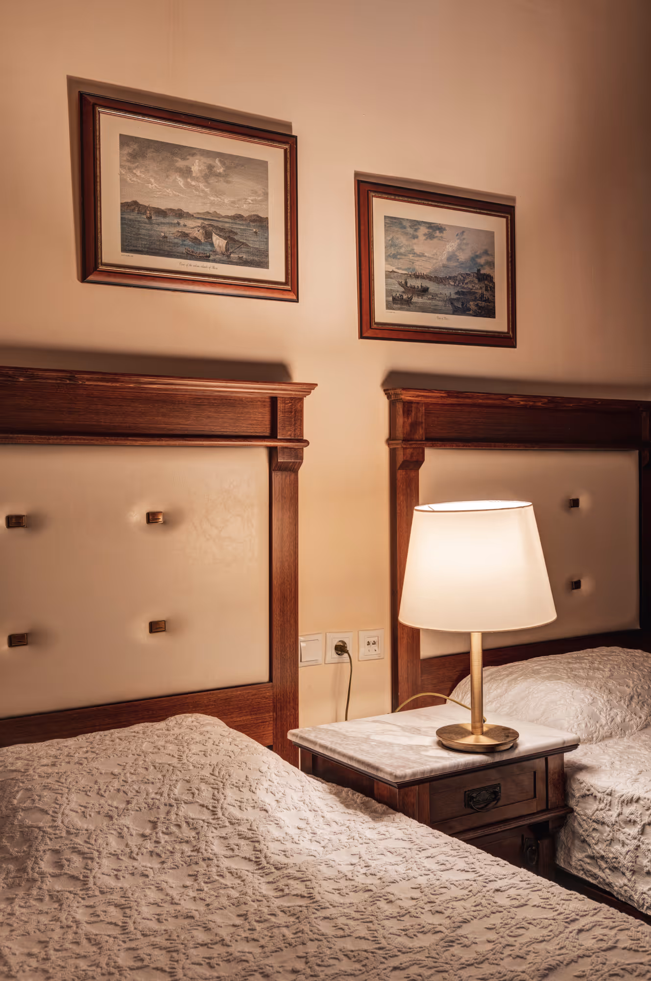 Two single beds with wooden headboards and cream tufted padding, separated by a nightstand with a lit table lamp, under framed artwork on the wall.