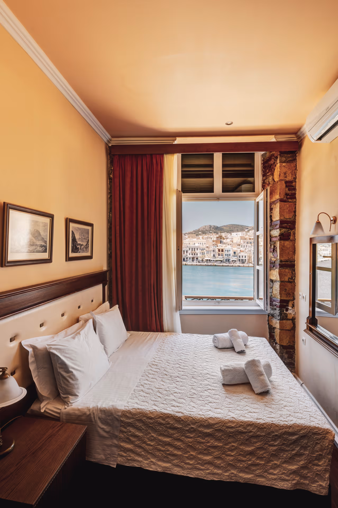 Cozy bedroom with a large bed, white pillows, folded towels, and an open window showing a waterfront town.