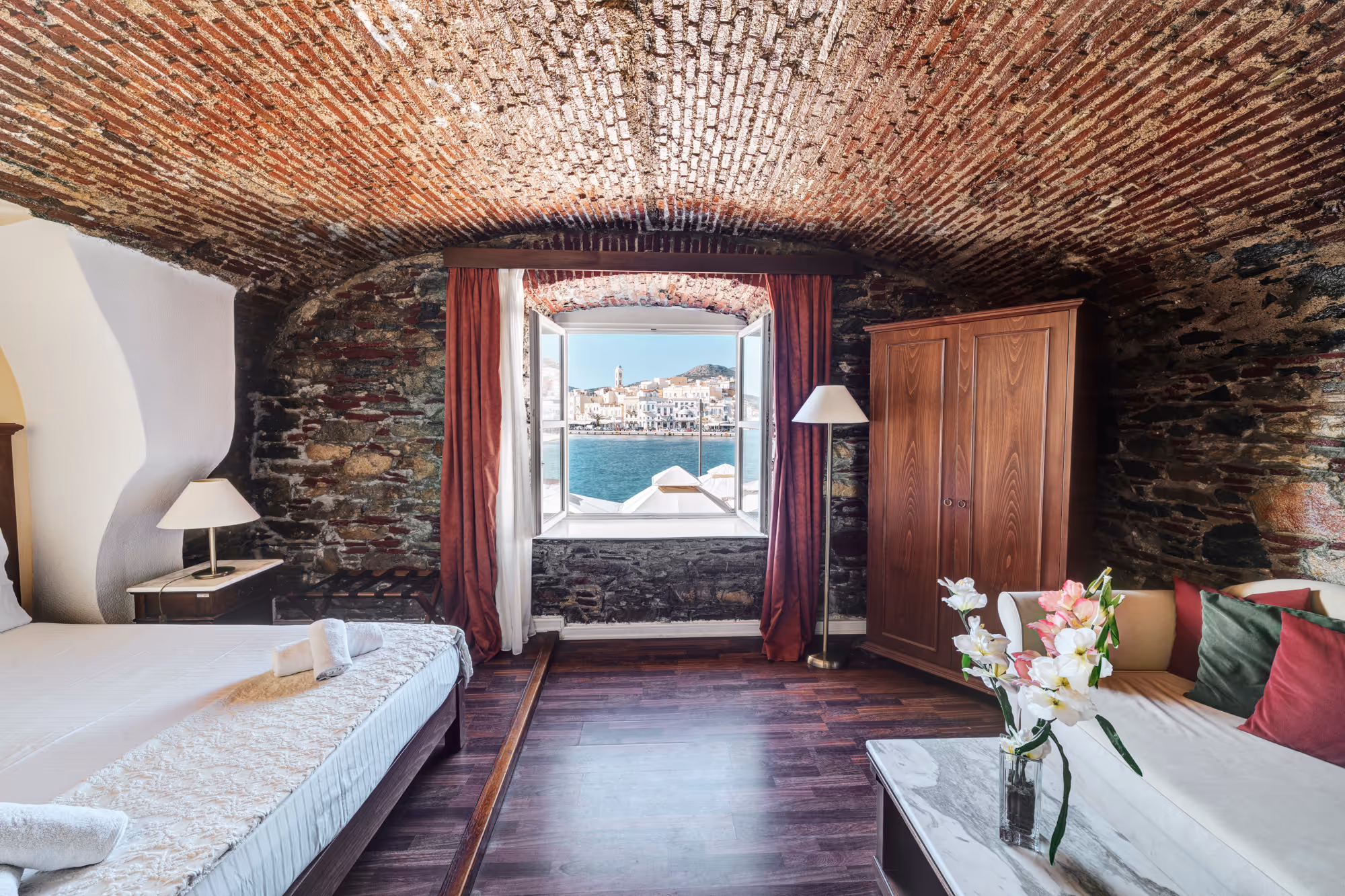Cozy room with arched brick ceiling, white bed, sofa with colorful pillows, wooden wardrobe, and window overlooking seaside town.