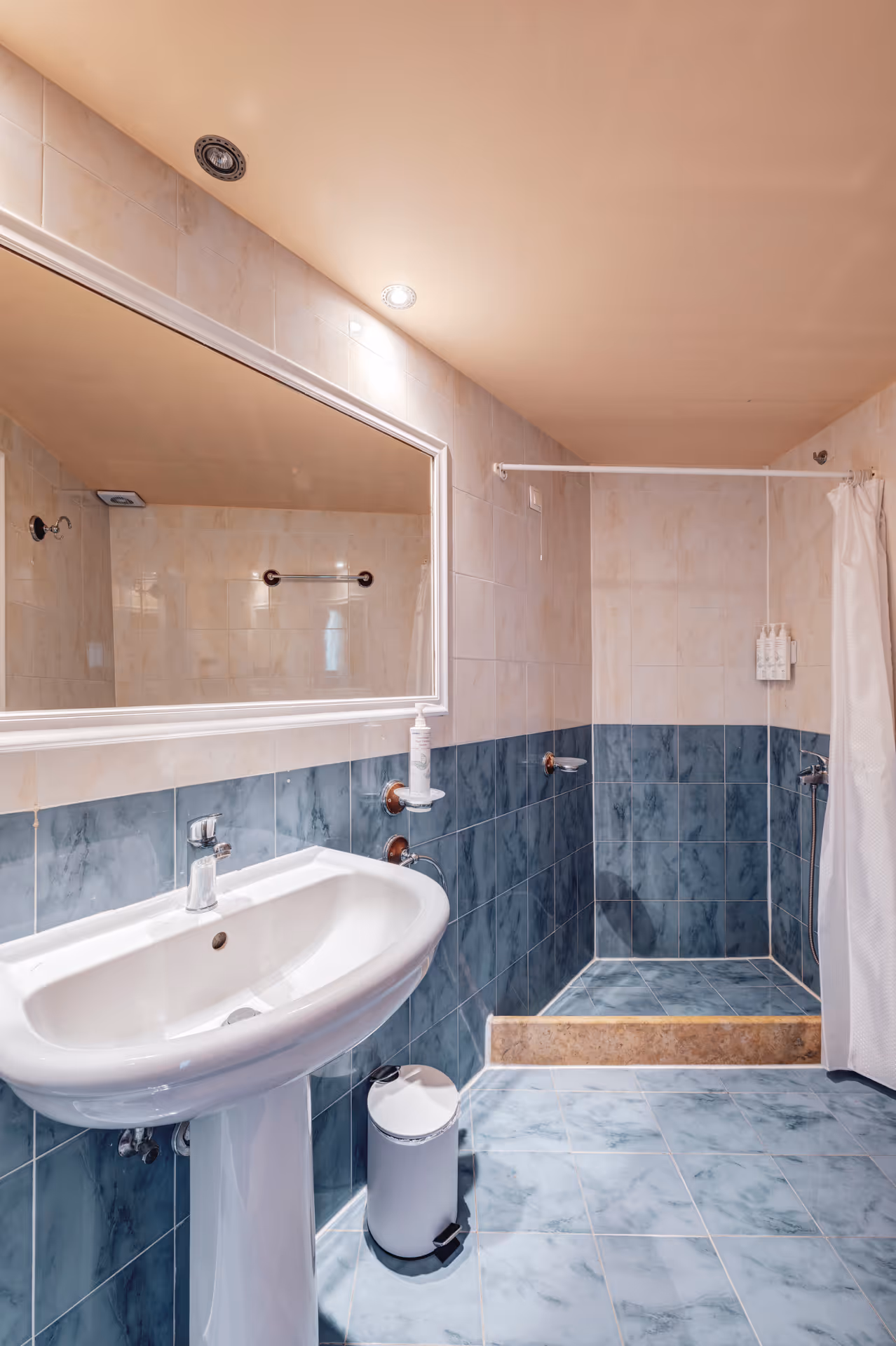 Bathroom with a white pedestal sink, blue and beige tiled walls and floor, a large mirror, and a shower area with a white curtain.