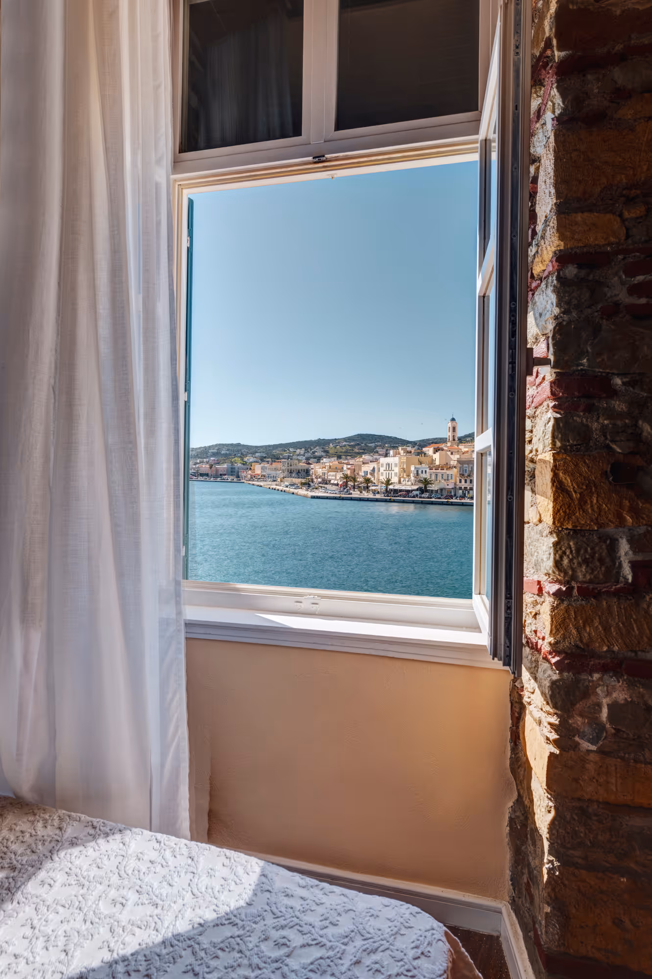 Open window in a room overlooking a seaside town with clear blue sky and calm water.
