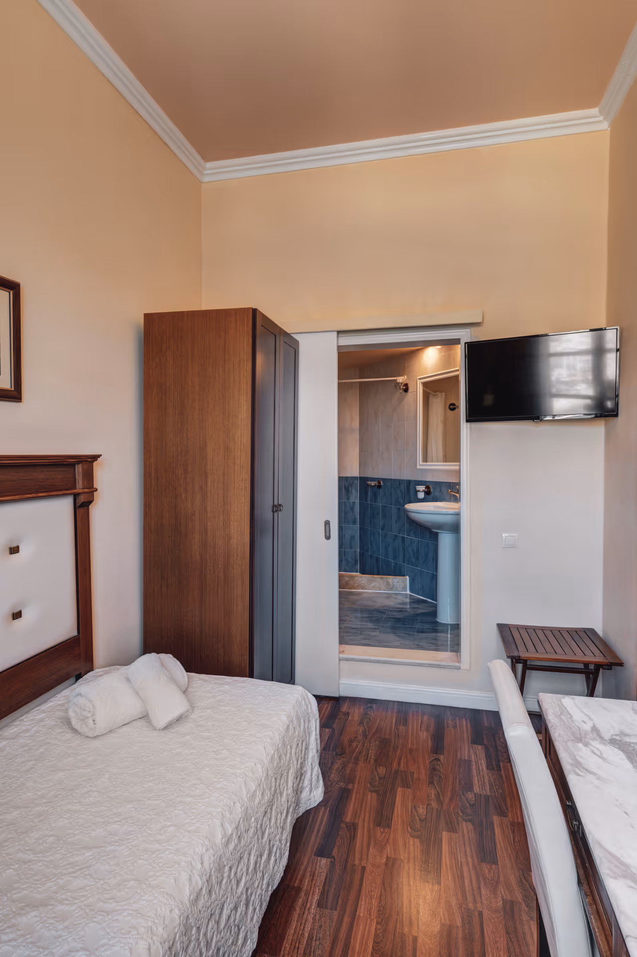 Small bedroom with a single bed covered in white bedding, a wooden wardrobe, a mounted flat-screen TV, and an en-suite bathroom visible through a sliding door.
