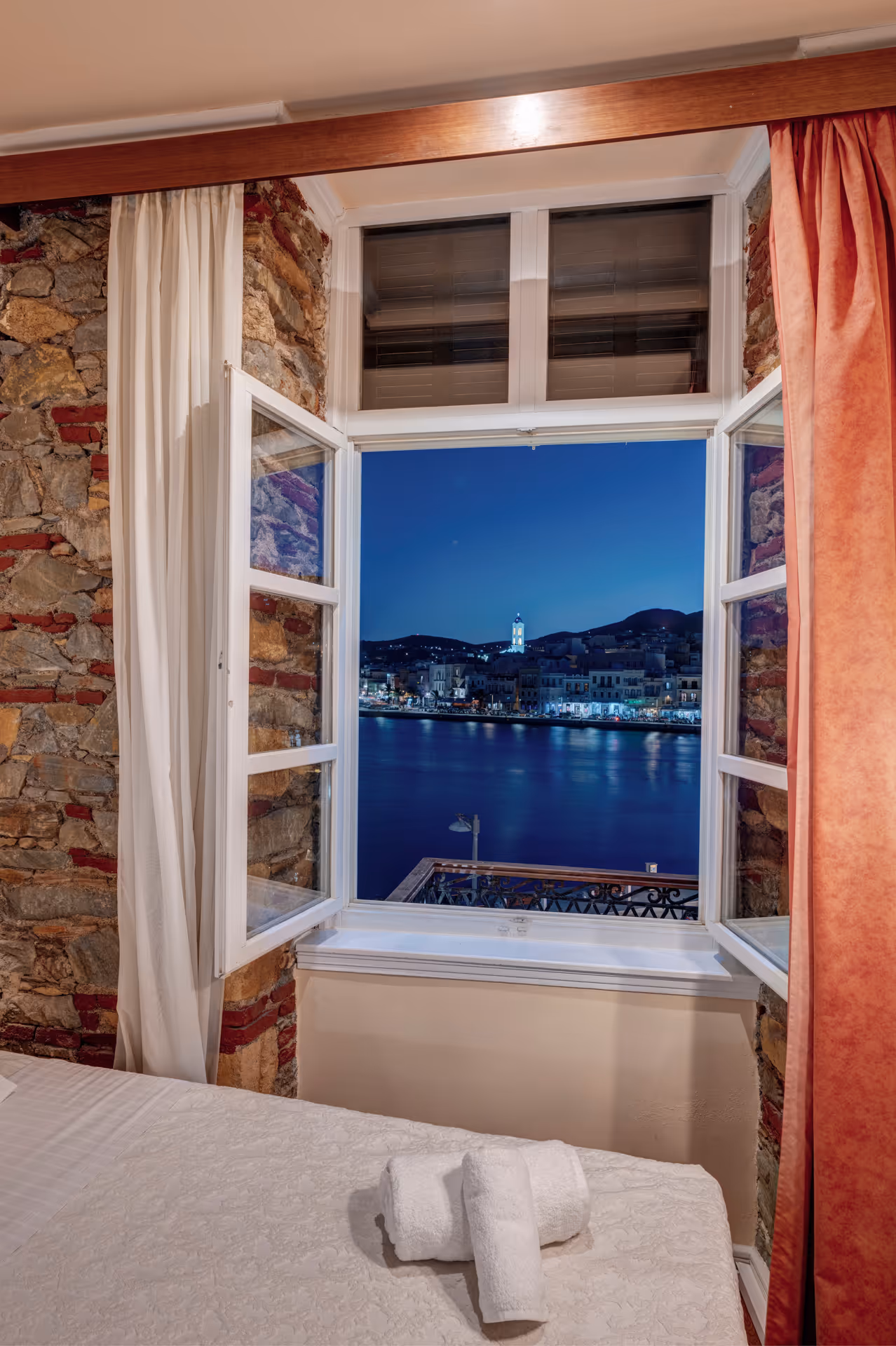 Open window in a rustic stone-walled room overlooking a calm waterfront town lit at night.