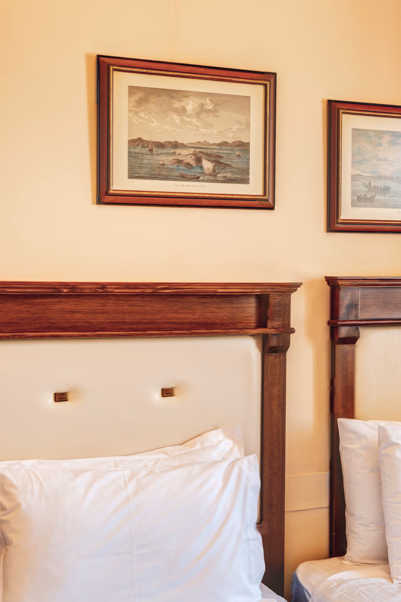 Part of two adjacent beds with wooden headboards and white pillows under framed seascape paintings on a beige wall.