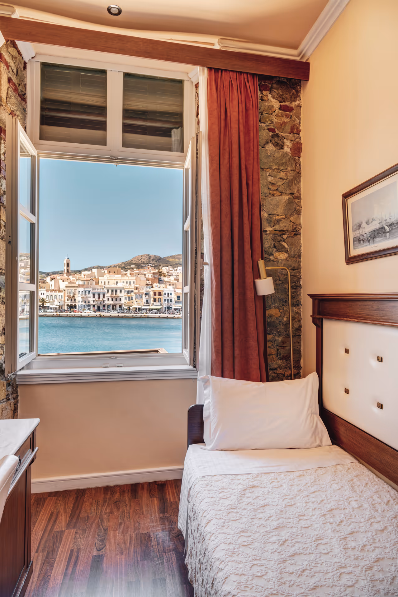 Cozy bedroom with single bed next to an open window showing a waterfront town and hillside under clear blue sky.
